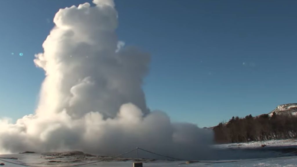 Iceland's Geysers: The Science Behind the Spectacle A powerful geyser erupting a tall column of steam and water into a clear blue sky, surrounded by a snow-covered landscape with trees and mountains in the background