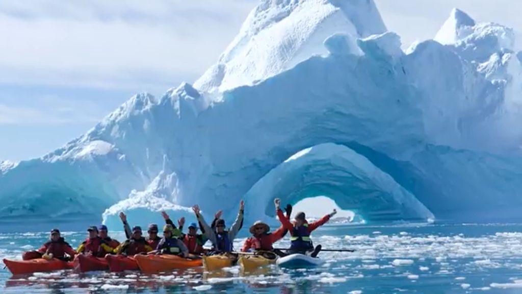 A group of kayakers in colorful red, orange, and yellow sea kayaks raising their arms in celebration, paddling in icy polar waters in front of a massive white and blue iceberg featuring a dramatic natural arch opening.