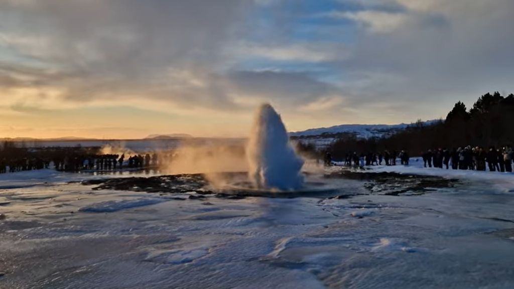 How to Travel Iceland Responsibly: A Guide for Conscious Explorers A geyser erupting a tall steam column from a snowy landscape at sunset, with a large crowd of tourists encircling it