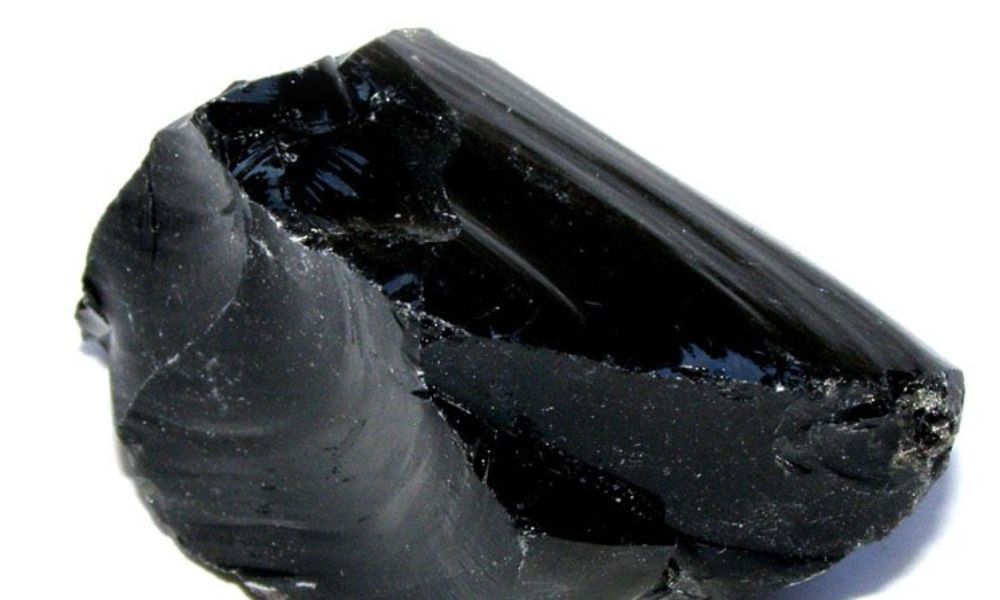 Close-up view of a black obsidian fragment on a neutral background, showing its smooth glassy surface and conchoidal fractures.