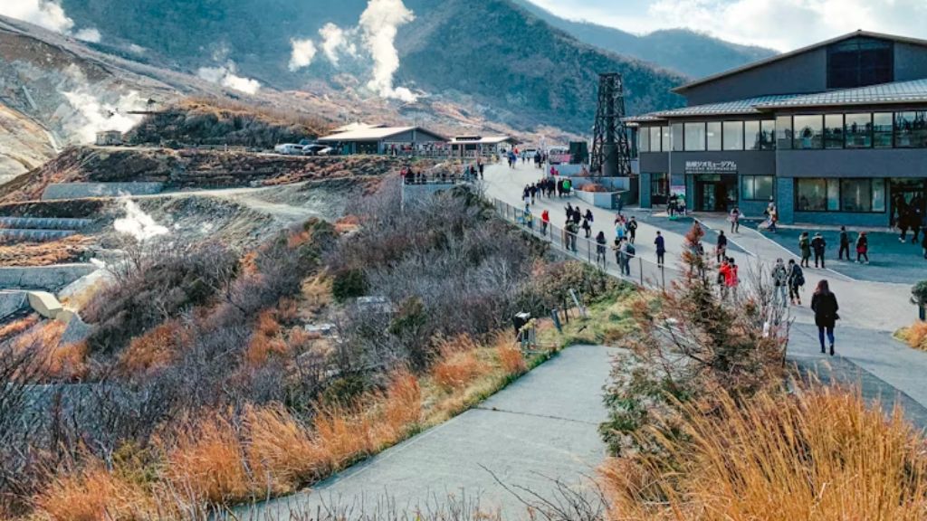 Visitors walking along a path through a volcanic valley with rising geothermal steam and surrounding mountains.