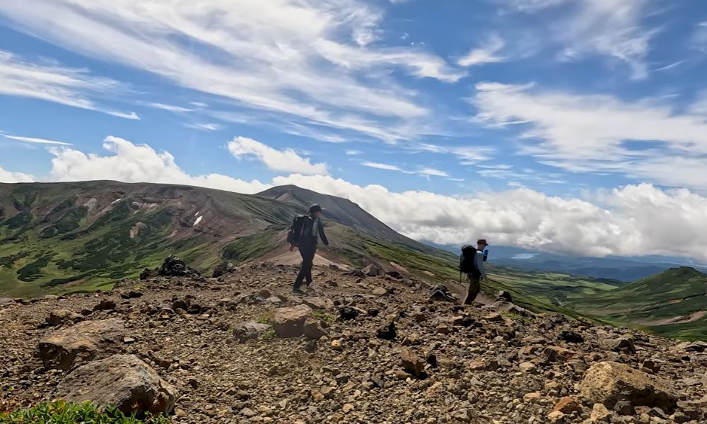 A hiker walking along a dark volcanic lava path in Japan, with rough basalt blocks, sparse vegetation, and distant hills under a partly cloudy sky.