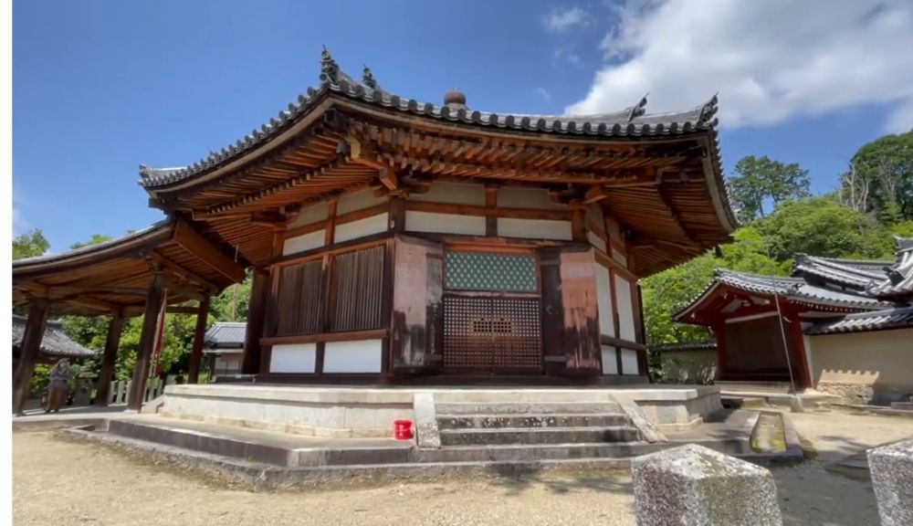 Traditional Japanese wooden temple with tiled curved roof, raised stone platform, and surrounding smaller buildings under a blue sky.