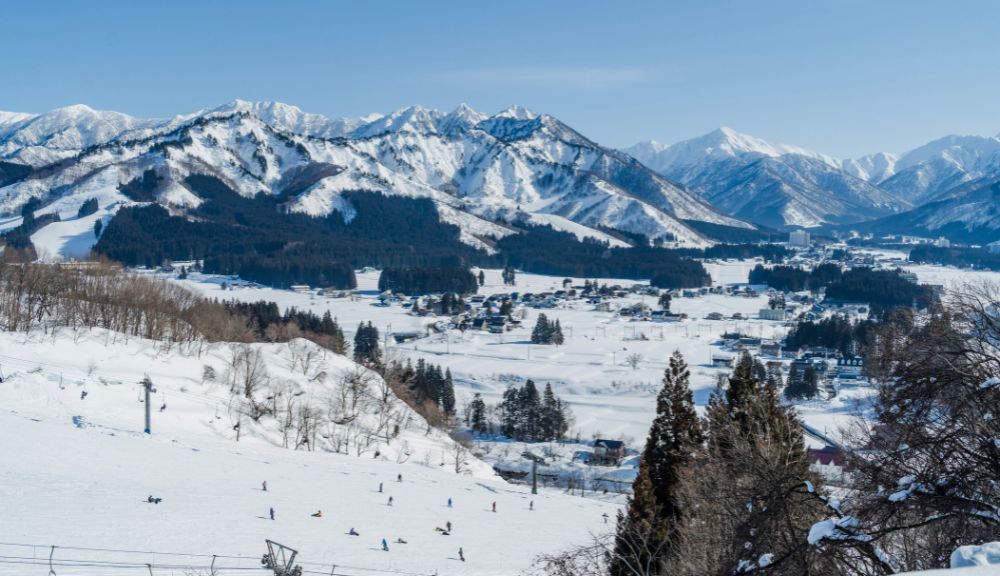Snowy ski resort with skiers on open slopes, a small town in the middle ground, and tall snow‑covered mountains rising under a clear blue sky.