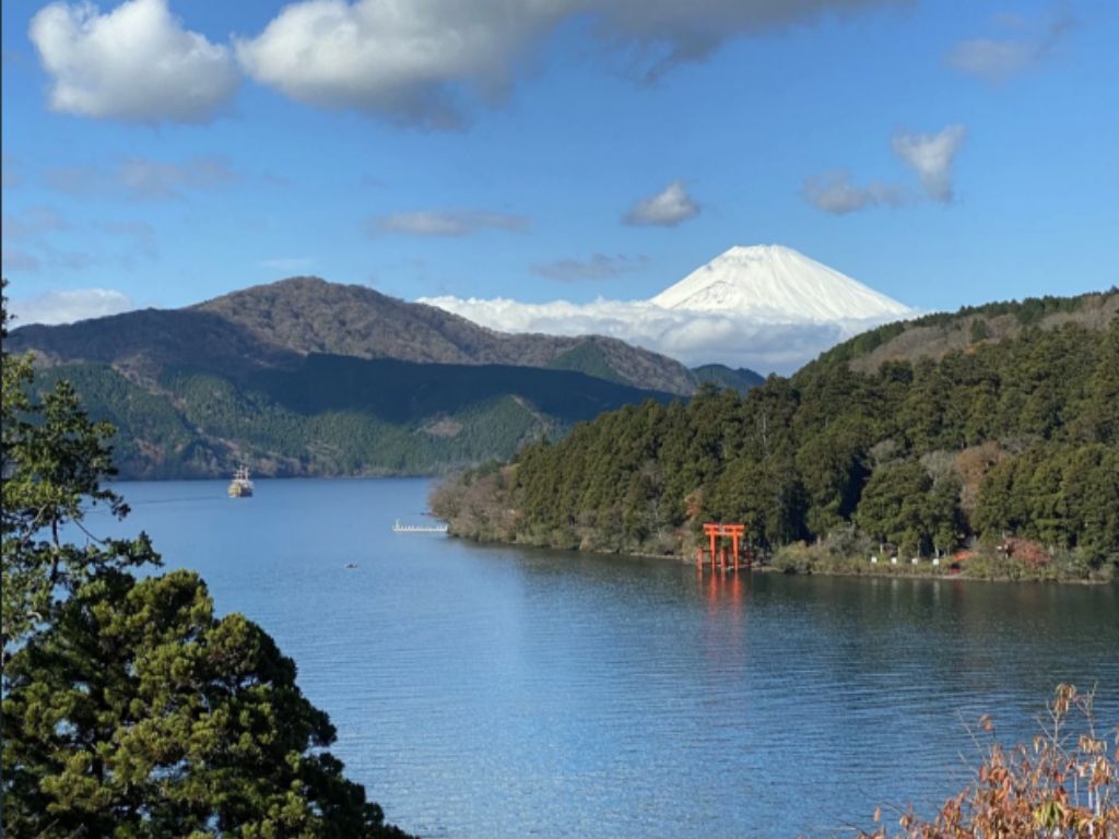 Snow-capped Mt. Fuji rising above forested hills, with Lake Ashi and a red torii gate in the foreground