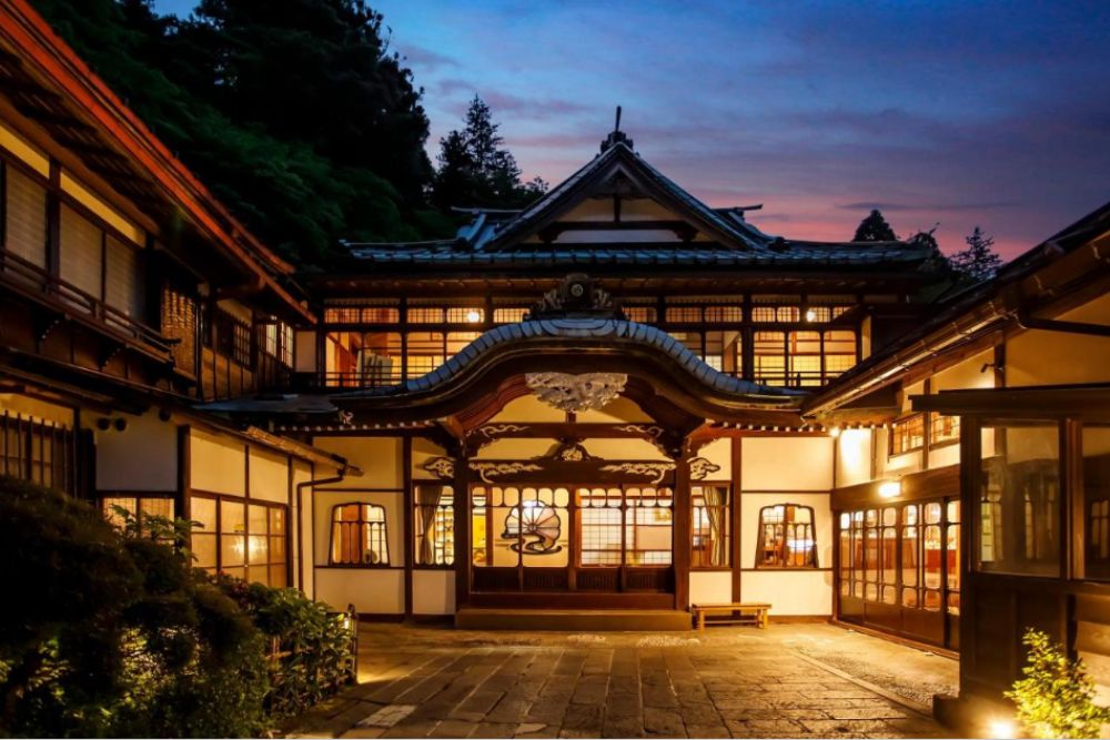 Exterior of a traditional Japanese ryokan illuminated by warm evening light, with wooden façades and soft lantern glow.