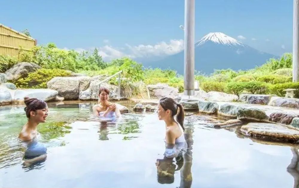 Three smiling women soak and chat in a rock-lined outdoor onsen pool, surrounded by green trees and large boulders, with a partial wooden pavilion structure visible and a snow-capped Mount Fuji visible in the distance under a bright blue sky.
