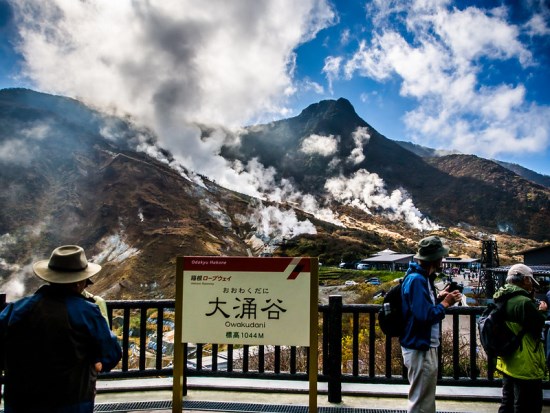 Tourists stand at a railed observation platform in front of a sign reading "大涌谷 Owakudani, elevation 1044M." Behind them, a rugged volcanic mountain emits thick white steam from multiple fumaroles under a partly cloudy blue sky.