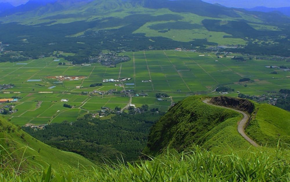 Cultivated fields and a quiet village on the floor of the Aso caldera, with Mount Nakadake fuming gently in the background.