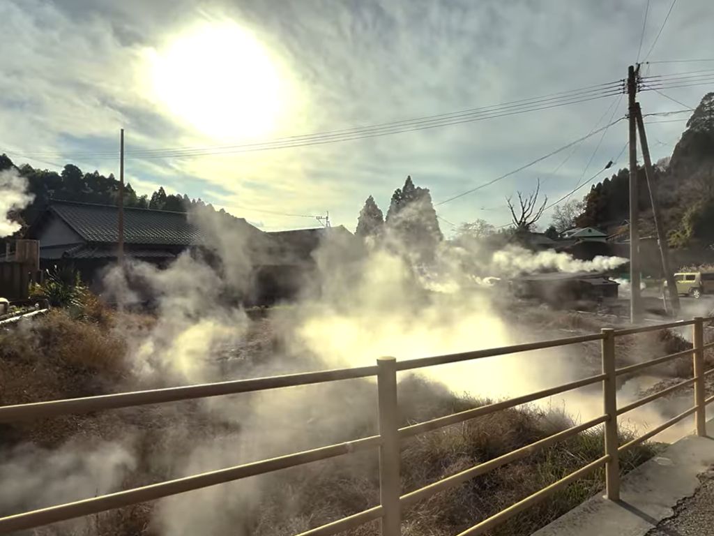 Steam emerging from geothermal vents near houses in a rural Japanese village, with forested hills in the background