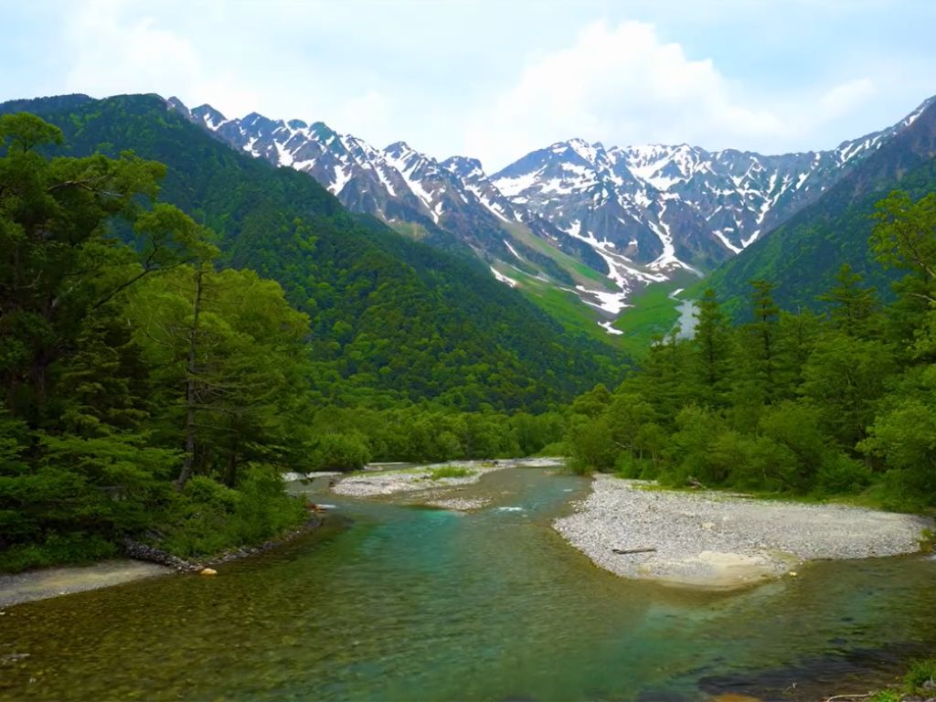 Mountain landscape of the Hotaka Range seen from Kamikōchi valley, with the Azusa River in the foreground and snow patches on grey granite peaks under a soft summer sky.