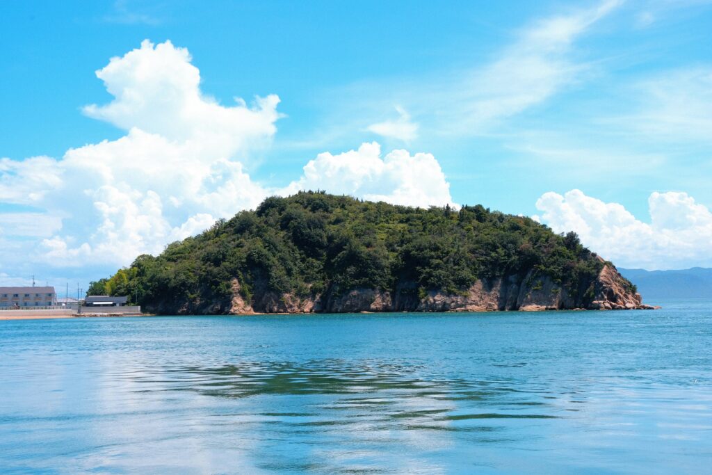 Forested granite islet rising from the calm turquoise waters of the Seto Inland Sea off Naoshima, Kagawa, Japan, under a blue summer sky.