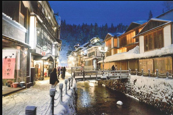 Snow‑covered onsen village at dusk, with warm light glowing from wooden inns and soft steam drifting above a stone canal, inspired by Kurokawa, Ginzan or Kinosaki.