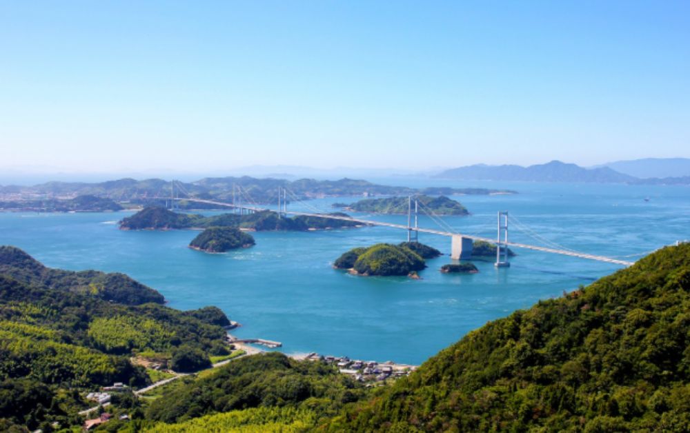 Panoramic view of the Seto Inland Sea from a hilltop, with forested granite islands and a long suspension bridge spanning the calm blue water.