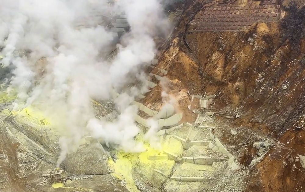 Yellow sulfur deposits and rising steam across a rocky volcanic hillside with erosion barriers.